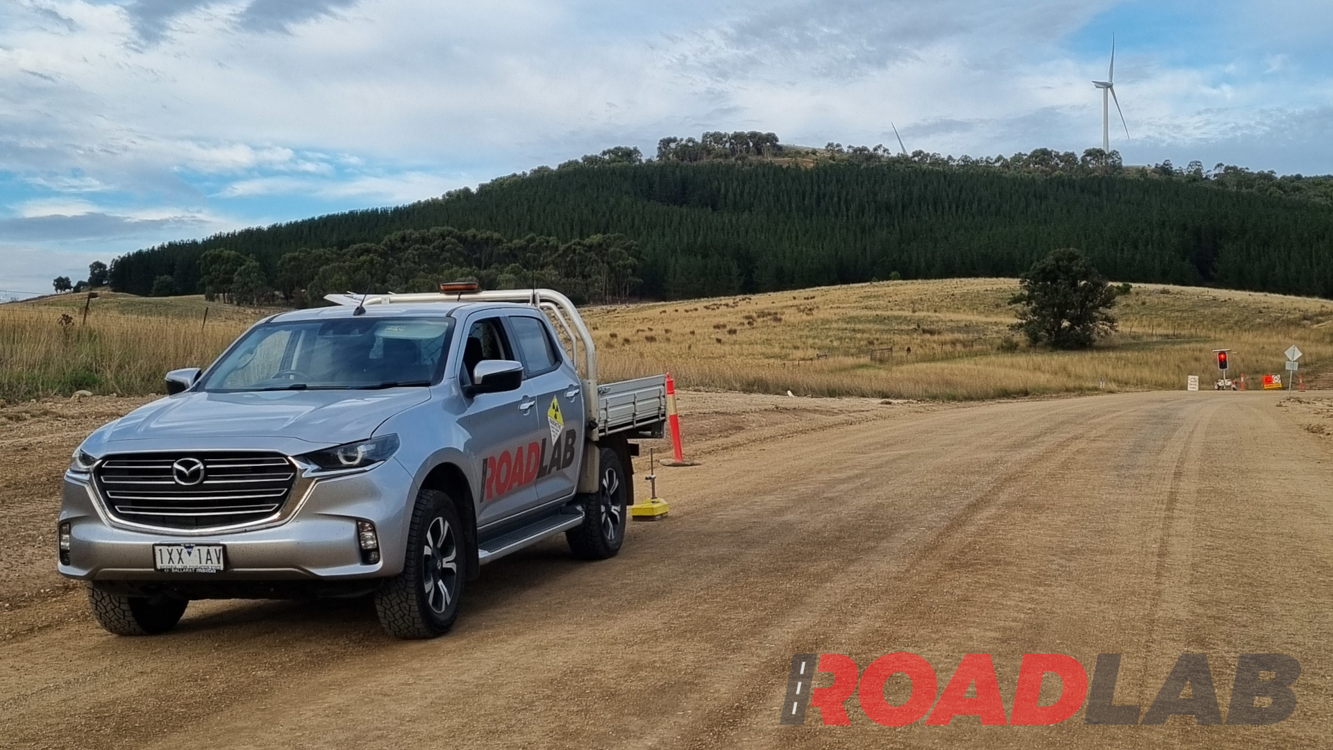 A rural road under construction, with a roadlab ute in the foreground and wind turbines on a hill in the distance.