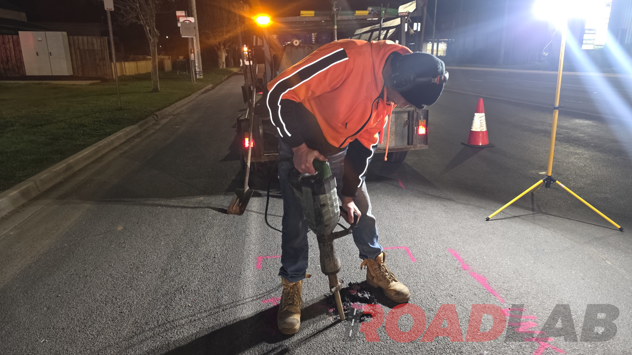 A person in high-visibility clothing using a jackhammer on an asphalt road at night.
